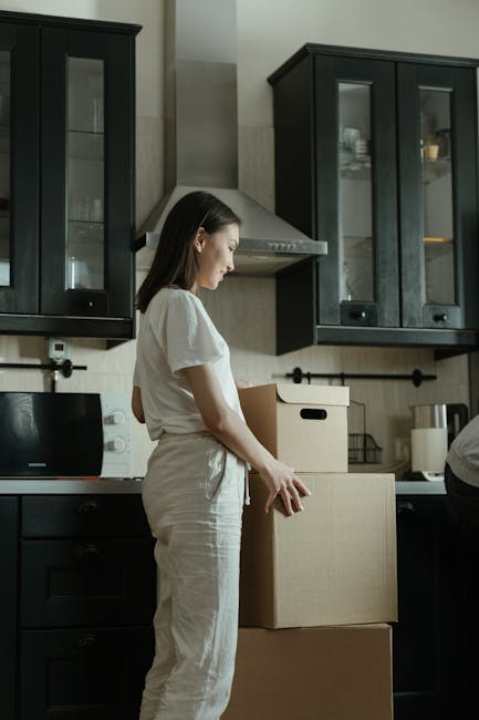 Close-up of a person packing house contents, with feet sticking out from inside an open cardboard box, wearing white socks. The scene is set indoors in a well-lit room with natural light coming throug