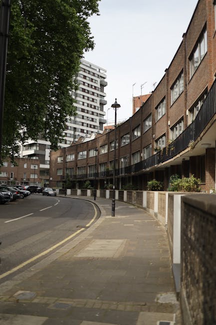A wide view of a residential street outside a multi-storey curved apartment building constructed with brick, featuring black metal balconies and large windows. The pavement is lined with small concret