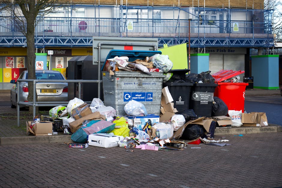 A row of four large wheelie bins placed on a cobblestone pavement outside a building with a pinkish-orange facade. The first bin on the left is dark green with a green label, the second is yellow with