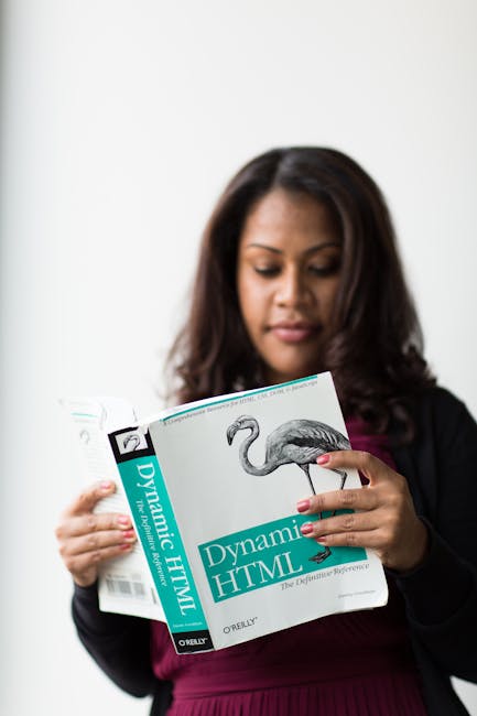 A woman with long dark hair, wearing a black blazer over a maroon top, is standing indoors against a plain white wall. She is holding a textbook titled 'Dynamic HTML' by O'Reilly in her left hand, wit